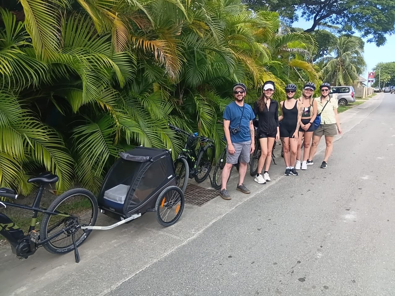 Group of six cyclists wearing helmets posing with bikes on a sunny street beside tropical plants and parked cars ahead.