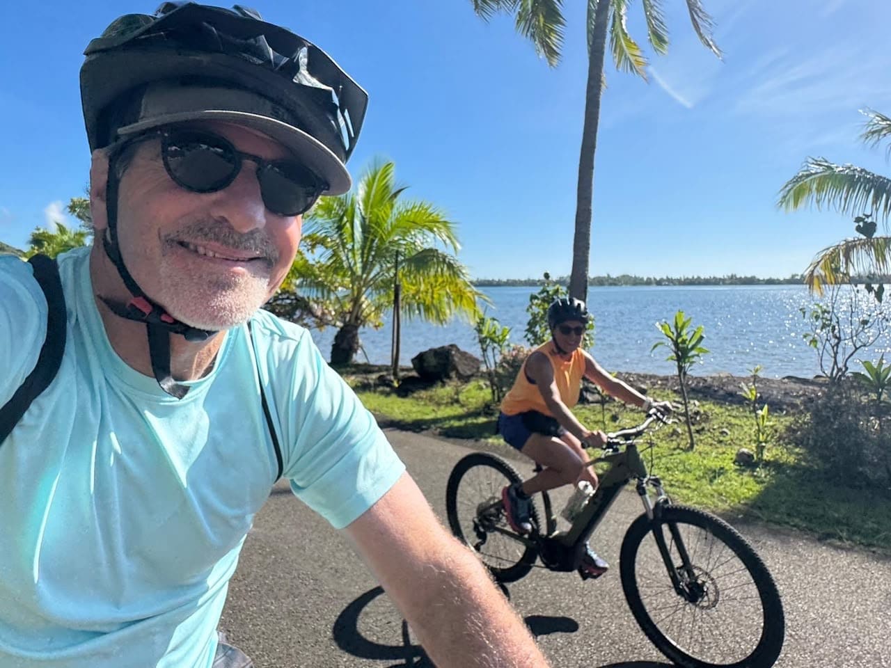 Smiling man wearing a helmet and sunglasses takes a selfie while riding a bike on a sunny waterfront path, with another cyclist in the background and palm trees nearby.